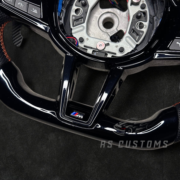 RS CUSTOMS BMW G-Series custom steering wheel with piano black trim, perforated leather grips, and orange stitching. Features LED display.